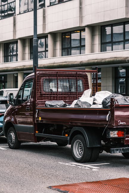 A small burgundy flatbed truck parked on an urban street in front of a modern building with large rectangular windows and concrete exterior panels. The truck's bed is loaded with various types of waste materials, including large black and white plastic bags, cardboard, and miscellaneous rubbish items. A metal mesh barrier extends vertically from the truck's cab to the edge of the bed, providing support and separation. The vehicle's body is clean, with visible small wheels and a simple tail light arrangement at the rear. The scene is illuminated by natural daylight, highlighting the texture of the truck's metallic surface and the urban environment around it. The context suggests an independent rubbish collection service, such as Waste Removal Earls Court, handling on-site clearance or private waste disposal for commercial or residential premises in the area.