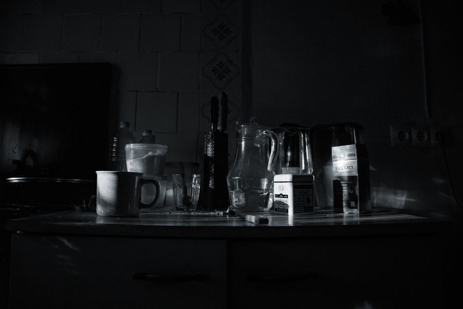 A dark, dimly lit kitchen countertop featuring various kitchen items and containers. In the center, there is a clear glass pitcher with a handle, positioned on a light-colored surface, along with a black kettle and a box of tea bags. To the left, a white ceramic mug with a smooth finish sits next to a small glass and a plastic water bottle. Behind these, a set of black utensils with long handles is held upright in a container. The background shows tiled walls with some visible grout lines and a partially visible window or light source casting subtle reflections on the shiny surfaces. To the right, there is a small box, possibly containing sugar or similar product, and a glass jug with a narrow spout. The overall scene appears cluttered and utilitarian, typical of a kitchen environment focused on daily use, with the lighting emphasizing textures and reflections on the glossy surfaces. A standard kitchen drawer handle is partially visible at the bottom edge, and there are slight shadows cast by the items, contributing to a moody, understated atmosphere. This scene subtly relates to the concept of personal or private waste handling, as the objects could be part of everyday waste or recyclables awaiting disposal, aligning with independent rubbish collection services like Waste Removal Earls Court.