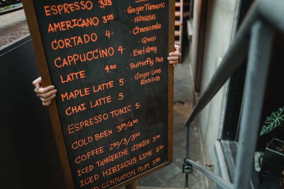 A person standing behind a blackboard-style menu board, holding its sides with both hands and partially visible. The menu displays various hot beverages such as espresso, americano, cortado, cappuccino, latte, maple latte, chai latte, espresso tonic, cold brew, iced tangerine ginger, iced hibiscus lime, and cinnamon plum in bright orange chalk-like lettering on a dark background. The menu includes prices ranging from £2.50 to £5 and features drink ingredients like ginger lime, hibiscus, chamomile, green, Earl Grey, butterfly pea, ginger turmeric, horseradish, and rooibos. The scene appears to be inside a café or coffee shop, with some visible elements of the interior environment, including a metal railing on the right and what seems to be a plant pot on the bottom right corner. The overall atmosphere is casual and inviting, suitable for a coffee outing, with natural lighting illuminating the scene. This image relates to the context of a customer ordering beverages, contrasting with waste management themes but highlighting the importance of proper disposal and waste segregation in café settings, which is relevant to rubbish removals or waste handling services provided by Waste Removal Earls Court, especially in commercial environments.