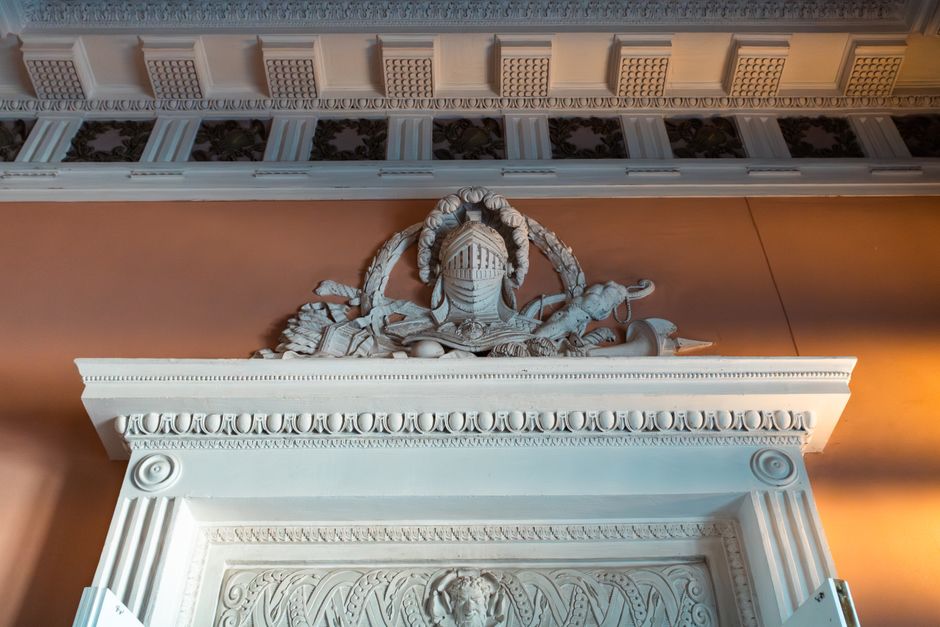 A detailed white plaster ornamental relief is mounted above a doorway within an interior setting. The decorative molding features classical design elements, including intricate beading, scroll patterns, and floral motifs, which are arranged symmetrically along the lintel. Above this molding, there is a sculptural composition consisting of a central figure wearing a helmet, flanked by various other sculpted objects that appear to be scrolls, ribbons, and possibly weapons or shields, all crafted with detailed textures. The background wall is painted in a warm, muted terracotta tone that contrasts with the white details, highlighting their craftsmanship. Natural light illuminates the scene, creating soft shadows that accentuate the three-dimensional quality of the carvings. The scene suggests an environment where historic interior architectural features are preserved or restored, indicative of a place such as a historic building, museum, or a property maintained for its decorative architectural heritage, with no visible rubbish or waste management activities present in the image.