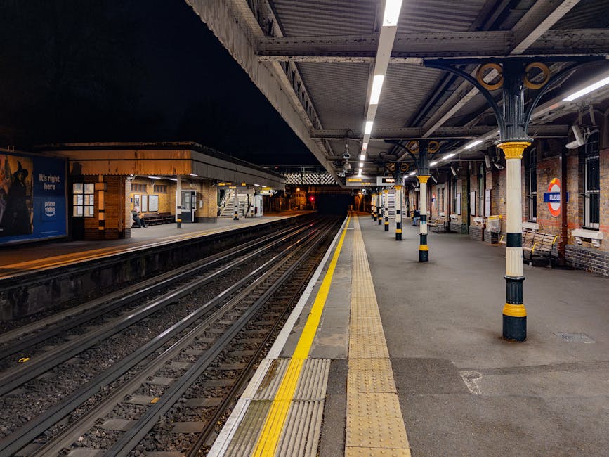 The image shows a quiet, well-lit underground railway station platform with a canopy supported by decorative columns painted black and yellow. The platform surface is made of concrete with tactile paving along the edge for safety, and a yellow line runs parallel to the train tracks, indicating a safe distance for passengers. Adjacent to the tracks are steel rails embedded in gravel, extending into the dark tunnel ahead. On the left side of the platform, there are benches, and a small brick building with windows and a staircase leading to an upper level is visible. To the right, a wall features a round London Underground sign with red, blue, and white colours, and several windows with brick surrounds. The station appears clean and empty, with a faint glow of artificial lighting highlighting the structured environment. This scene illustrative of London underground train stations, relevant to public transport and urban infrastructure, with no visible rubbish or waste on the platform or tracks. Waste Removal Earls Court offers alternative waste handling, but no waste removal tools or waste materials are present in this image.