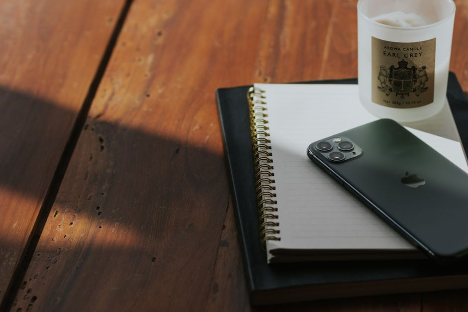 A close-up view of a wooden desk surface with visible grain and warm brown tones, partially illuminated by natural light casting soft shadows. On the desk, there is a black hardcover notebook with a smooth finish, placed beneath a white spiral-bound notepad with lined pages. Resting on top of the notepad is a modern black smartphone with a sleek, glossy finish and three camera lenses on the back. To the right, a white scented candle in a glass container labeled 'AROMA CANDLE EARL GREY' with a decorative emblem is partially visible. The scene depicts a tidy, organized workspace suitable for personal or professional use, with a subtle connection to waste management services emphasizing organized disposal or administrative planning related to rubbish removal, fitting within a context of private waste handling or administrative tasks associated with rubbish clearance. The visual atmosphere is calm and clean, highlighting details such as the textures of the materials and the arrangement of objects for a professional, neutral tone.