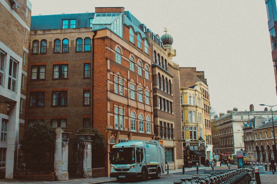 A street scene in an urban area featuring a collection of historic and modern buildings with various architectural styles, including brick facades and decorative window designs. The foreground shows a small waste collection vehicle parked on the pavement, with a compact, grey body and a black crew cab, likely used for local rubbish collection or private waste removal. Behind the vehicle, the street is lined with bollards and metal bike racks, with some bicycles secured to them. The background displays a mixture of multi-storey buildings, including a landmark building with a dome and a weather vane on top, as well as contemporary glass-fronted structures. The lighting suggests a cloudy or overcast day, providing diffuse natural light across the scene. This image relates to waste management services, illustrating the urban setting where independent rubbish collection and alternative waste handling options, such as private waste removal, are visible in the context of surrounding historic buildings and city streets.