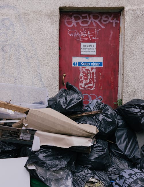 A large pile of black rubbish bags and discarded cardboard and wooden materials stacked in front of a small red door set into a textured, off-white building wall. The rubbish bags appear full and tightly packed, with some items partially visible beneath, including white foam and flattened cardboard. The red door features graffiti tags in white and pink spray paint near the top, a white sign indicating 'No Money' and 'Keep Clear' in blue and white, and a small, worn metal handle on the left side. The environment suggests an urban alleyway or backstreet area where waste has been temporarily deposited, potentially for collection or disposal by a private waste removal service such as Waste Removal Earls Court, offering an alternative to conventional rubbish collection. The scene is illuminated evenly, with no visible shadows, highlighting the accumulation of waste materials in a somewhat disorderly arrangement close to the door, emphasizing the importance of effective rubbish management in busy city settings.
